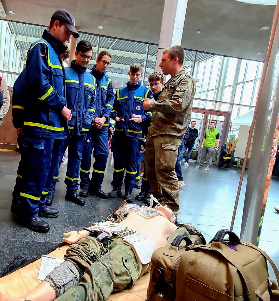 THW-Junghelfer stehen zusammen mit einem Bundeswehrsoldaten neben einer medizinischen Übungspuppe.