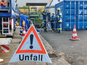 Im Vordergrund ein Schild: Unfall. Im Hintergrund Junghelfer mit Beleuchtung