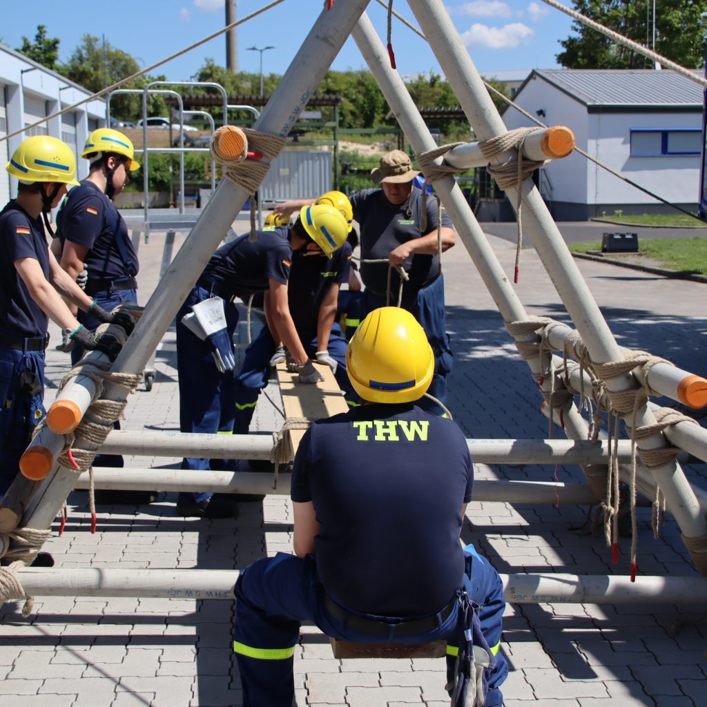 Zwei Junghelfer auf einer Wippe, die aus vielen Rundhölzern und Arbeitsleinen gebaut wurde. Andere Junghelfer sehen zu.